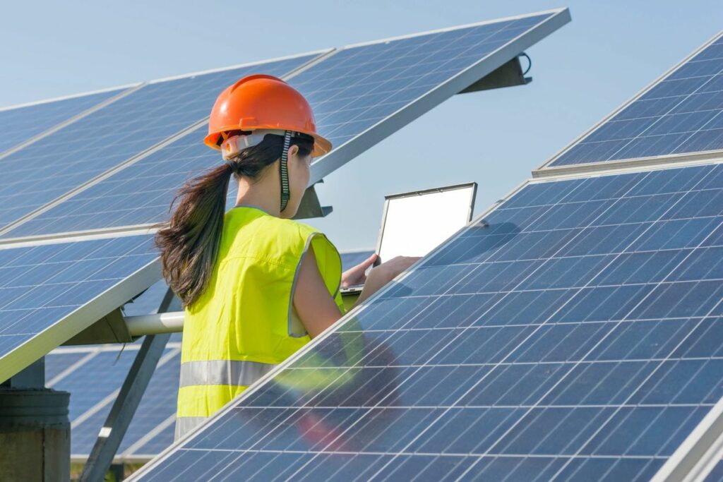 Worker in safety gear inspecting solar panels with a tablet.